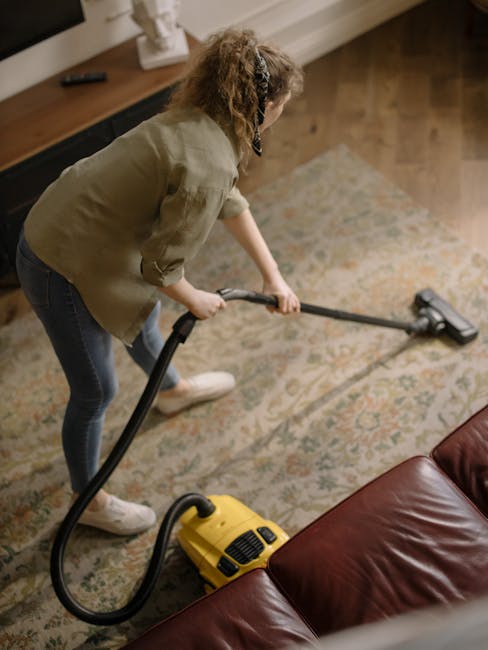 A person wearing full protective cleaning gear, including a white disposable coverall, mask, gloves, and shoe covers, is performing surface cleaning on a beige carpet using a professional vacuum or steam cleaning machine. The room has light-colored walls, a large mirror, and a wooden shelving unit with decorative items. Potted plants are visible in the background, and there is natural light illuminating the space. The scene depicts deep cleaning and sanitisation of a residential carpet, with attention to hygiene, carried out by Wimbledon Carpet Cleaning, a professional cleaning service in Wimbledon. This image highlights the importance of thorough domestic cleaning to maintain a healthy living environment.