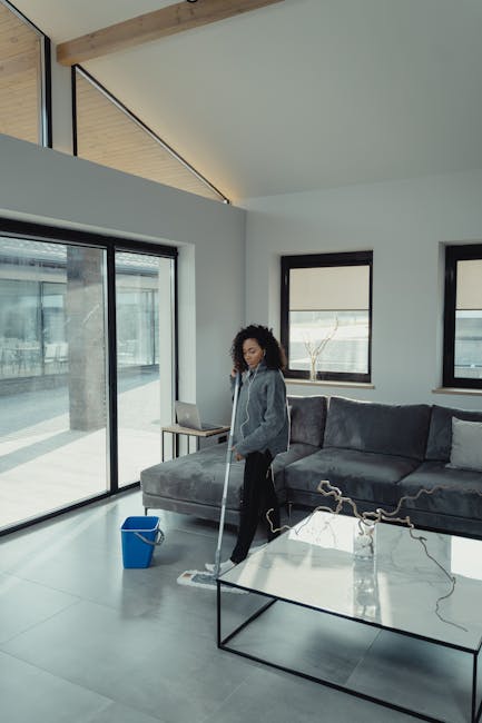 A woman with curly hair dressed in a grey hoodie and black pants is mopping a large, light grey tiled floor in a modern, bright living room. The room features white walls, two windows with black frames, and a black-framed glass sliding door that leads outside. A blue mop bucket is positioned nearby on the floor. There is a dark grey sectional sofa with cushion seating and a glass coffee table with black metal legs in the foreground. Natural light fills the room, highlighting the clean, polished surface of the floor. The setting exemplifies professional domestic cleaning service by Wimbledon Carpet Cleaning, maintaining a hygienic and well-presented living space.