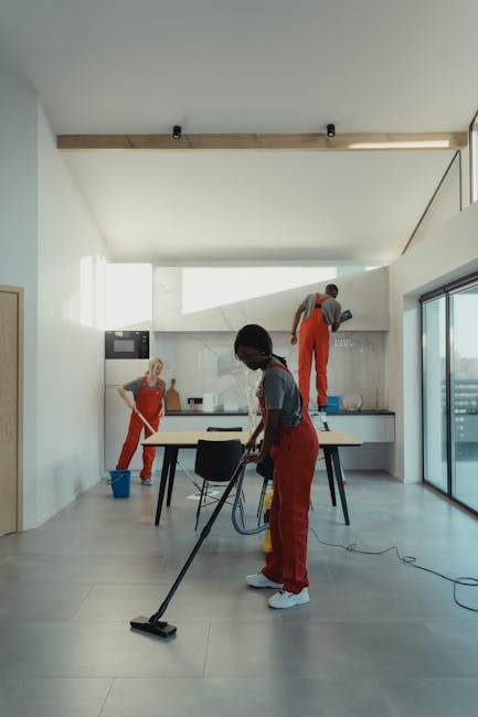 A team of three professional cleaners working in a modern, well-lit kitchen area with a large window and sliding glass door leading outside. One cleaner, dressed in red overalls and a grey shirt, is vacuuming the light grey tiled floor, focused on surface cleaning. In the background, another cleaner in red overalls is standing on a step ladder, reaching the ceiling panel or high wall area, possibly performing sanitisation or deep cleaning. The third cleaner, also in red overalls and a grey shirt, is wiping the kitchen counter with a cloth, ensuring surface hygiene. The kitchen features white walls, a sleek white countertop, a stainless steel oven, and minimalistic cabinets, all appearing clean and well-maintained. Natural light floods the space, enhancing the cleanliness and shiny appearance of the surfaces. Wimbledon Carpet Cleaning offers comprehensive domestic cleaning services, ensuring hygiene and cleanliness in residential spaces.