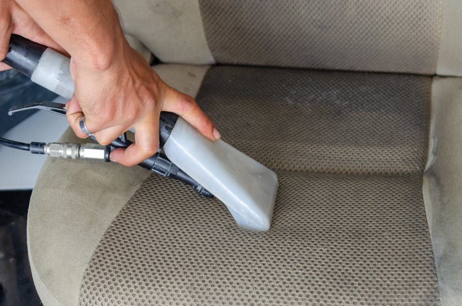 A professional cleaner's hand holding a handheld steam cleaning device, actively deep cleaning a beige upholstered sofa cushion with a textured pattern, in a well-lit room. The sofa's surface appears damp and in the process of being sanitised, with visible moisture and areas of cleaner or water residue. The scene highlights a focus on surface cleaning and hygiene, demonstrating domestic cleaning techniques used by Wimbledon Carpet Cleaning for upholstery refreshment, with the presence of velvet or microfiber materials and the use of specialized cleaning tools visible.